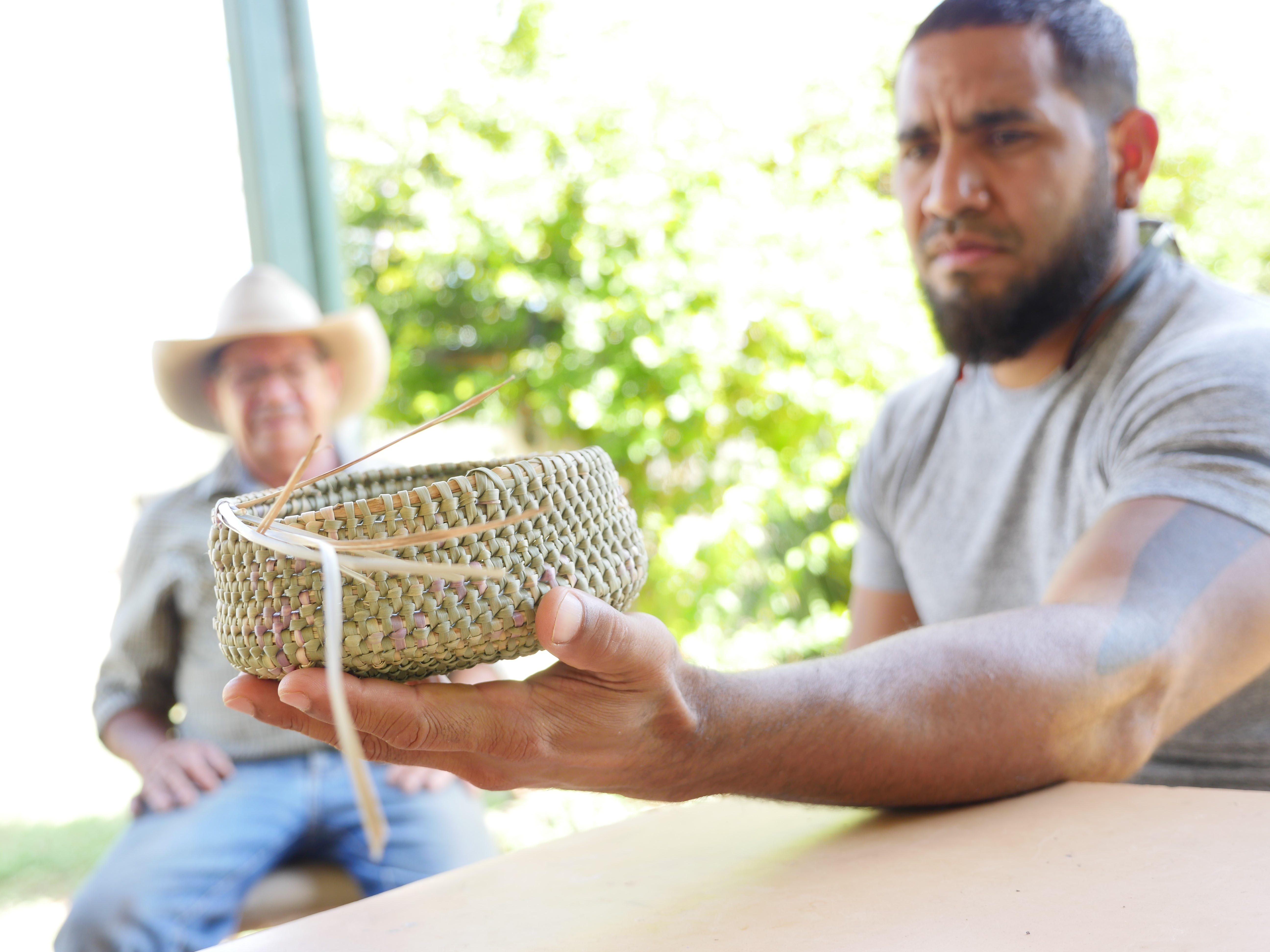 young indigenous man holding woven basket with older indigenous man sitting in the background, both men are out of focus